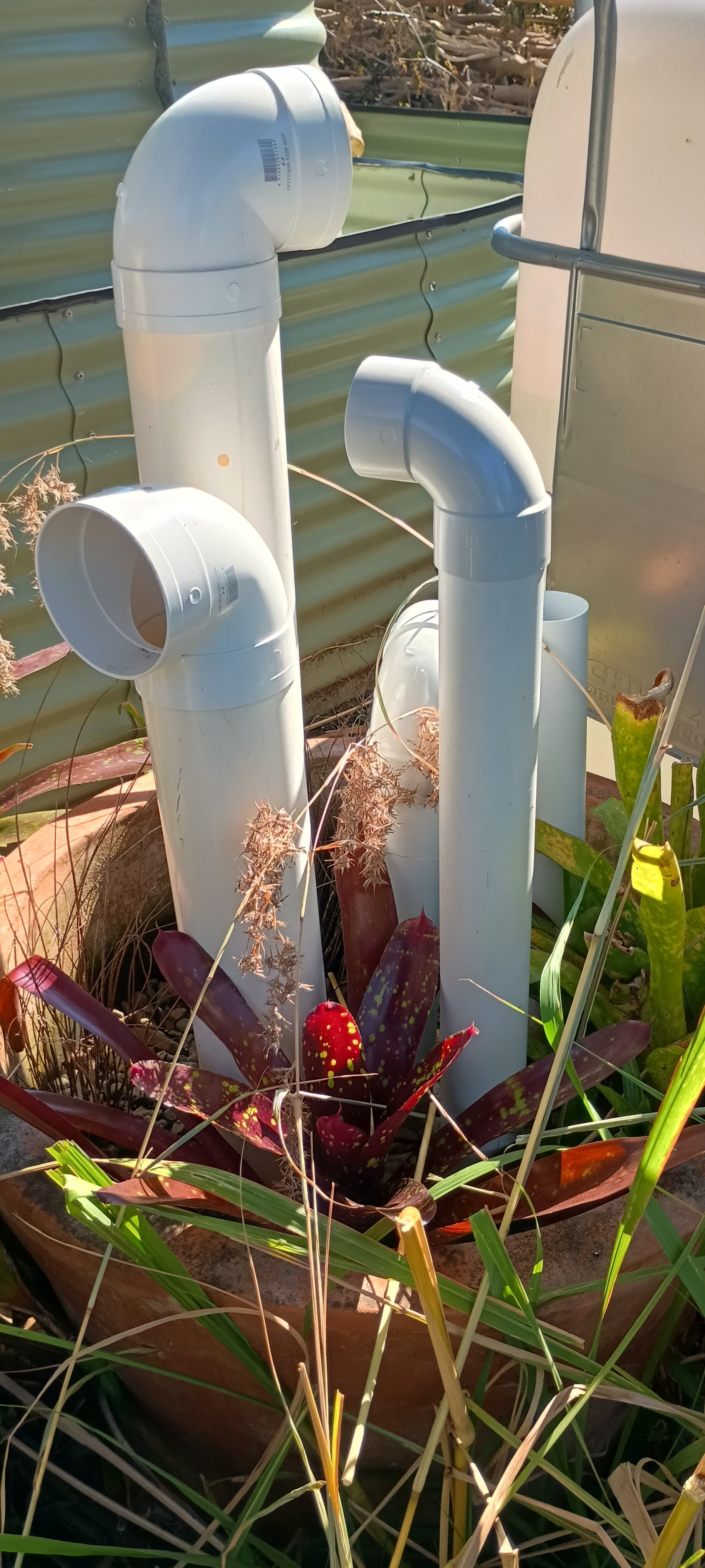 Two frog hotels made from white PVC elbow and pipe sections mounted on corrugated steel, surrounded by bromeliads and garden plants