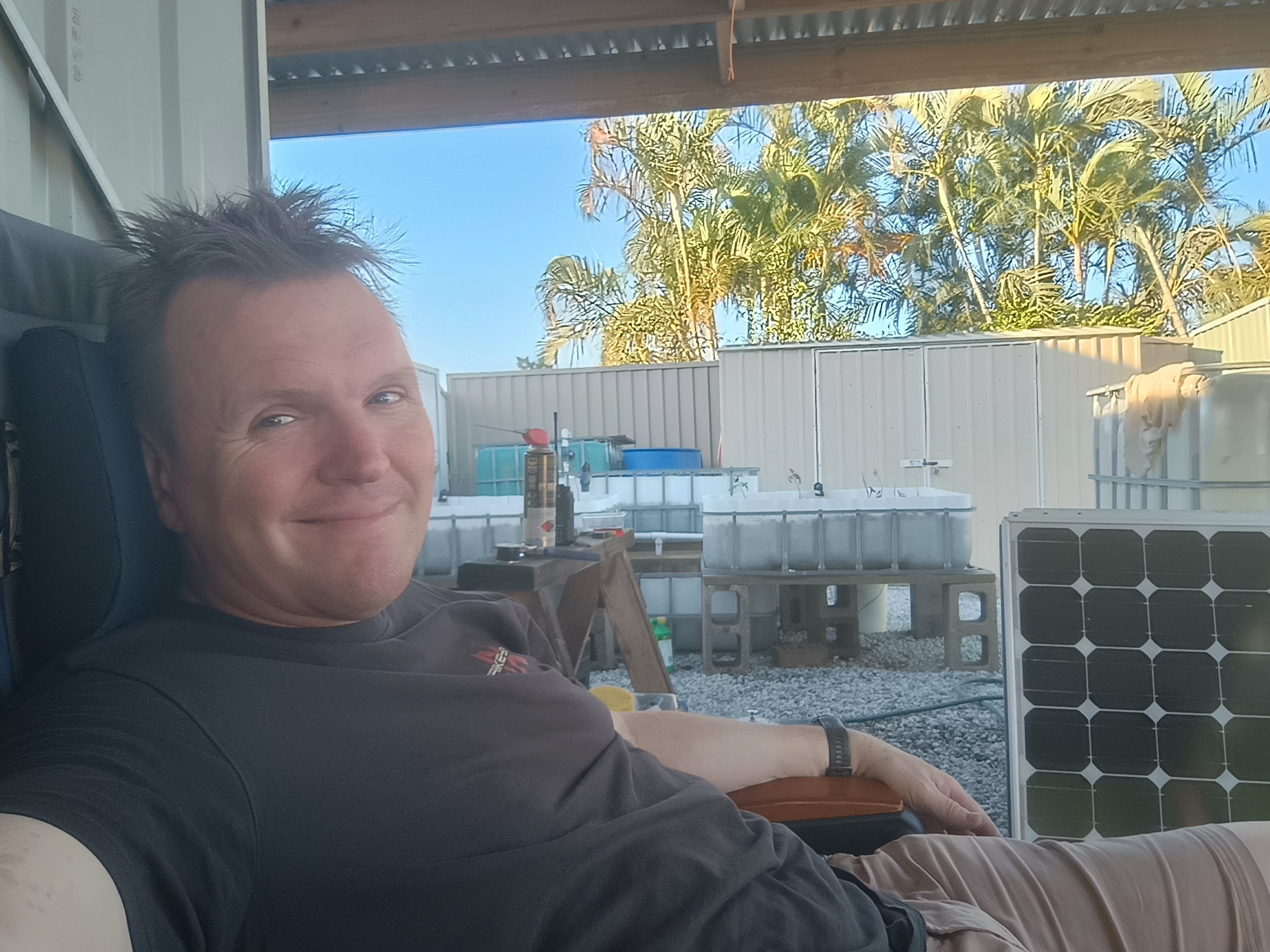 Steve Dalton relaxing in a chair under the shanty at the end of the day, with IBC tanks and a solar panel visible behind him
