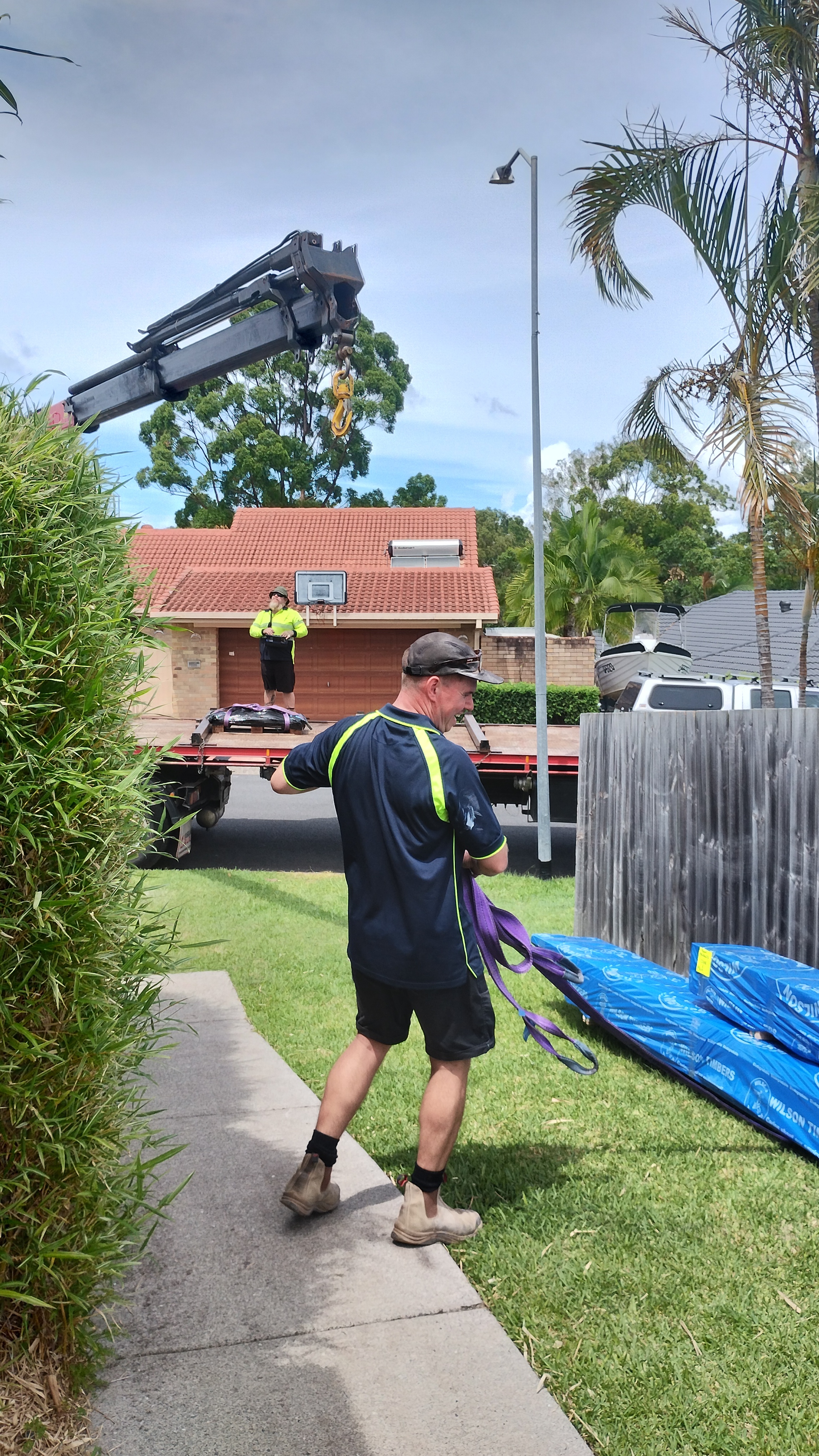Builder in hi-vis gear guiding lifting straps on a crane truck during timber delivery to a suburban backyard