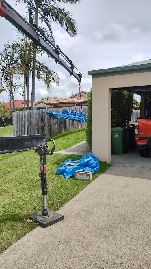 Crane truck delivering timber to a suburban backyard, with wrapped bundles being lifted over the fence