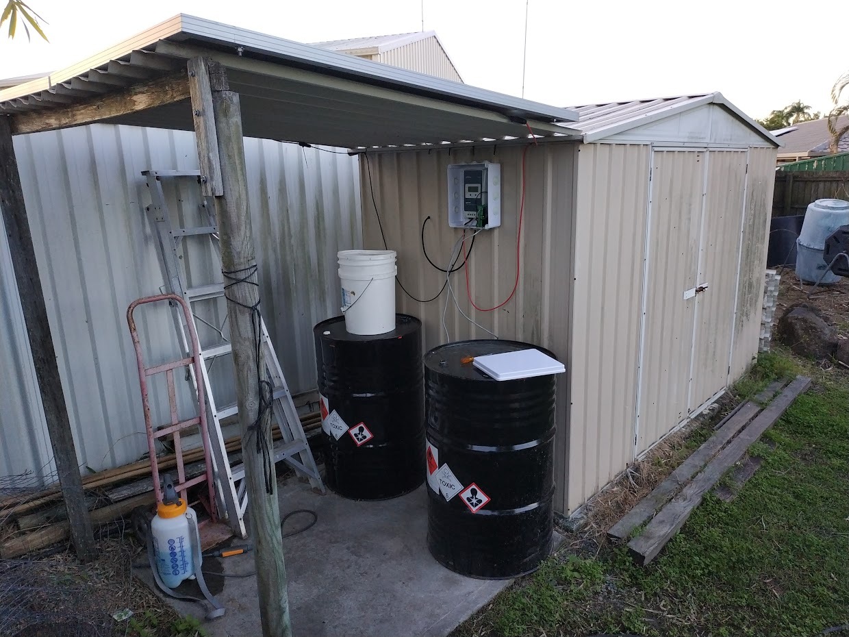 A homemade micro-shanty lean-to beside a garden shed, with two solar panels forming the roof, two repurposed black drums underneath, and an MPPT charge controller wired on the shed wall