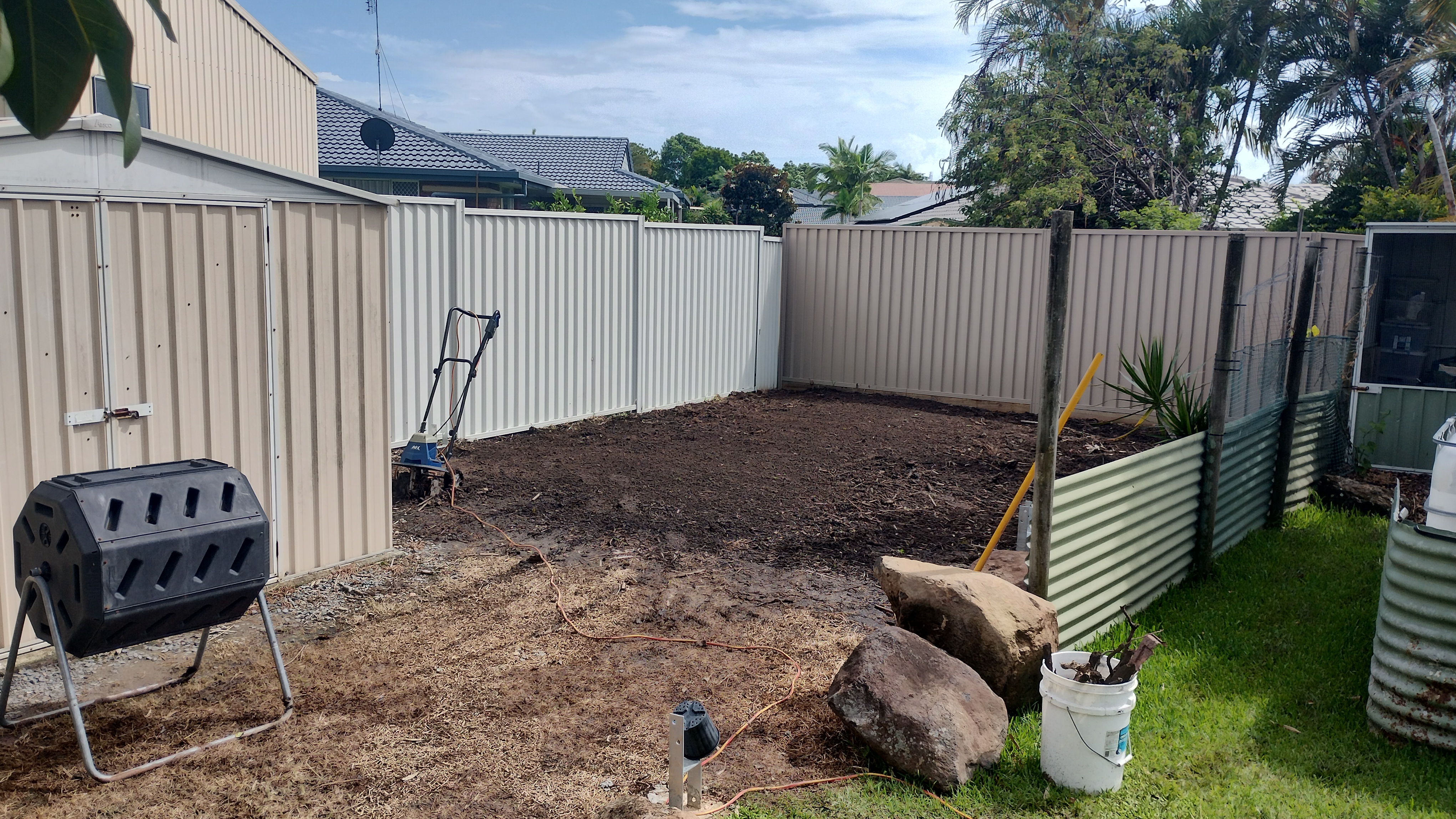 Backyard area cleared and cultivated, with a compost tumbler, large rocks, and corrugated steel raised garden beds -- previously compacted ground from chickens and bamboo